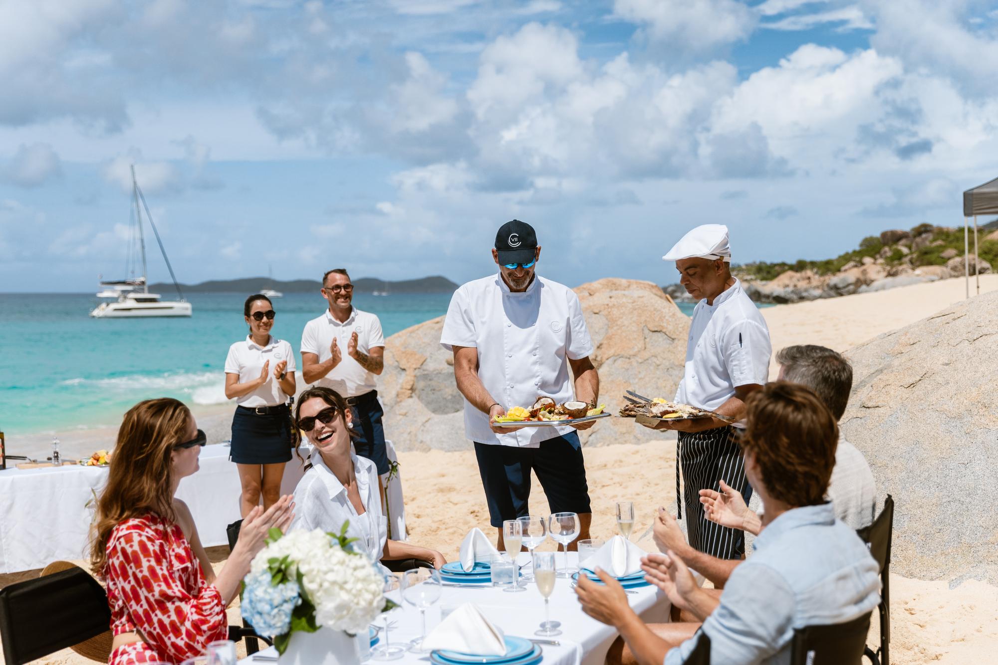 The CARINTHIA VII yacht's crew serving a delicious lunch on the beach, showcasing an elegant setup with gourmet food and attentive service for a luxurious outdoor dining experience. | CARINTHIA VII yacht, crew serving lunch, beach dining, luxury experience | Fraser Yachts