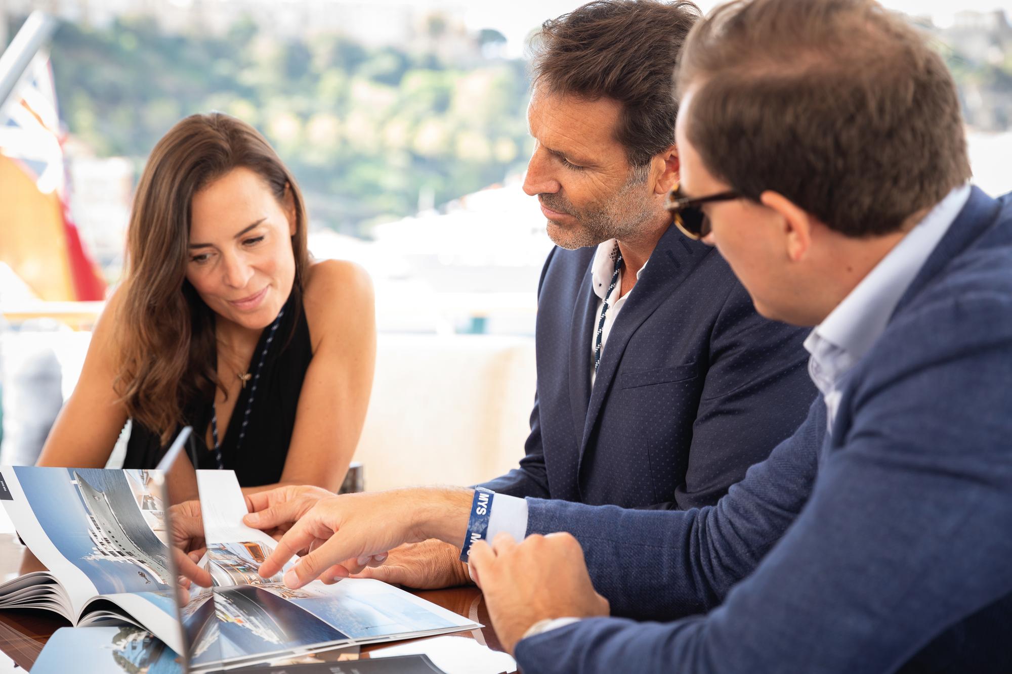 A build advisor discussing yacht options with clients, showcasing the latest designs in a magazine during a prospecting session. | yacht consulting, client meeting, yacht design magazine, build advisor | Fraser Yachts