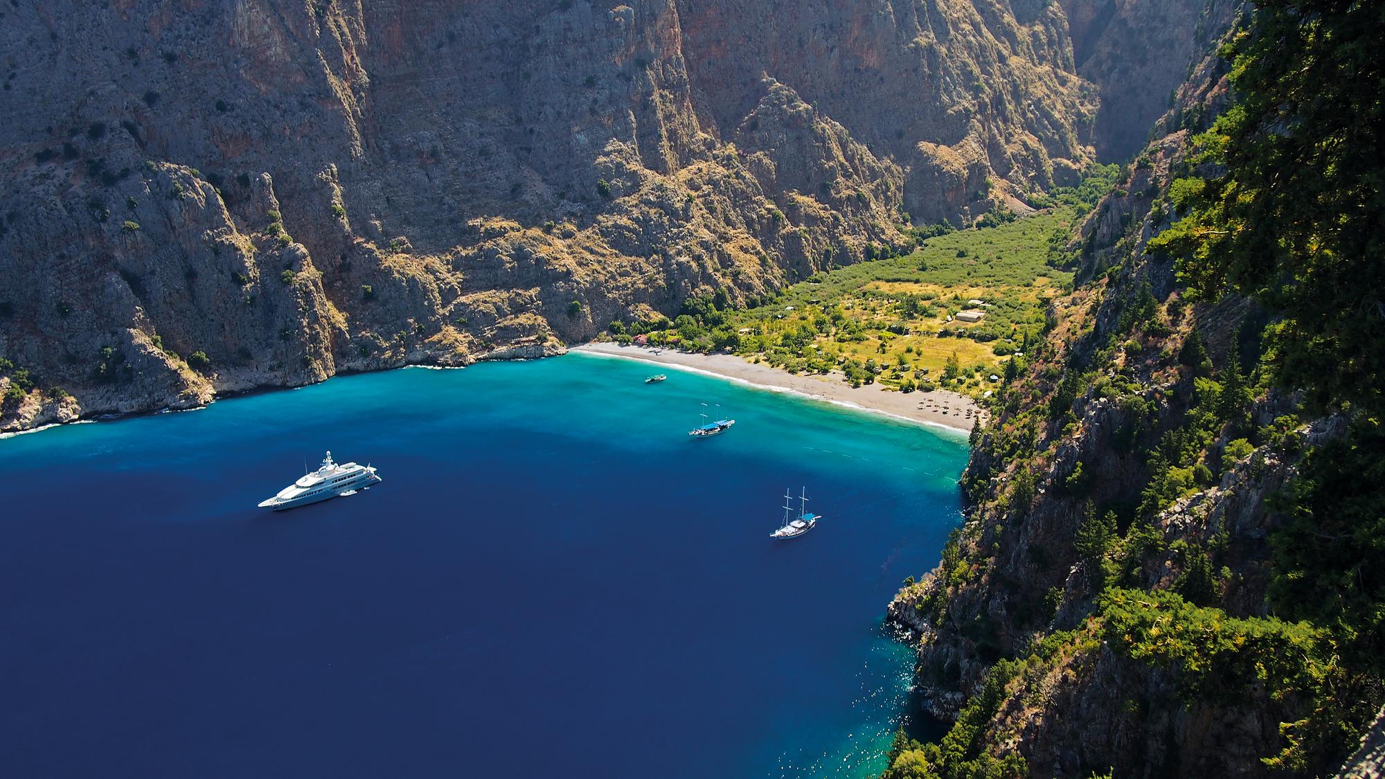 Embark on a captivating journey through the scenic beauty of Butterfly Valley in Turkey, where the sea meets the mountains, with Fraser Yachts. | Mediterranean, Turkey, Butterfly Valley, aerial view, sea, mountains, boats, yacht charter | Fraser