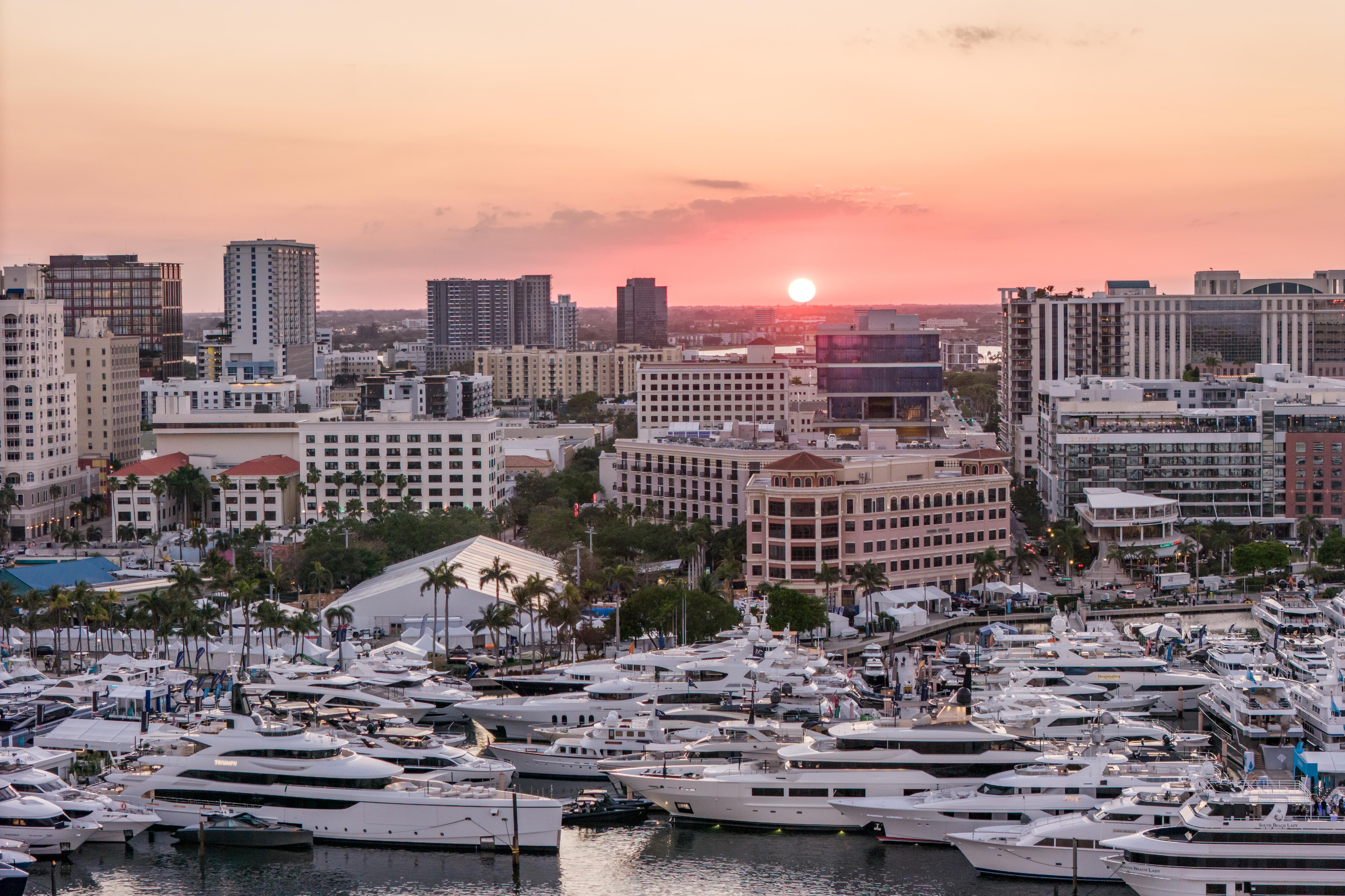 Luxury yachts docked at the Palm Beach International Boat Show, with a stunning city skyline and sunset in the background.