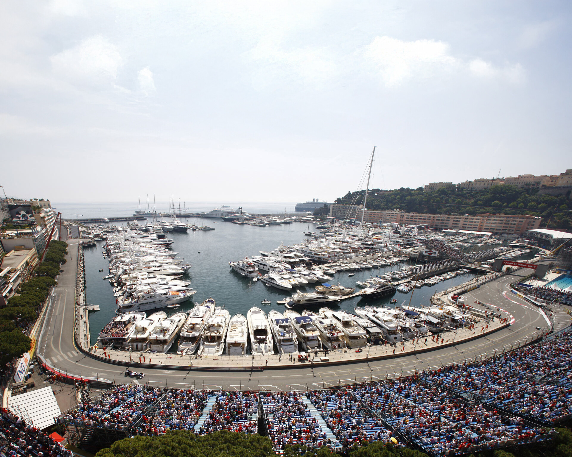 Aerial view of the Monaco Grand Prix circuit winding around Port Hercules, with luxury yachts, a packed crowd, and a Formula 1 car racing along the waterfront