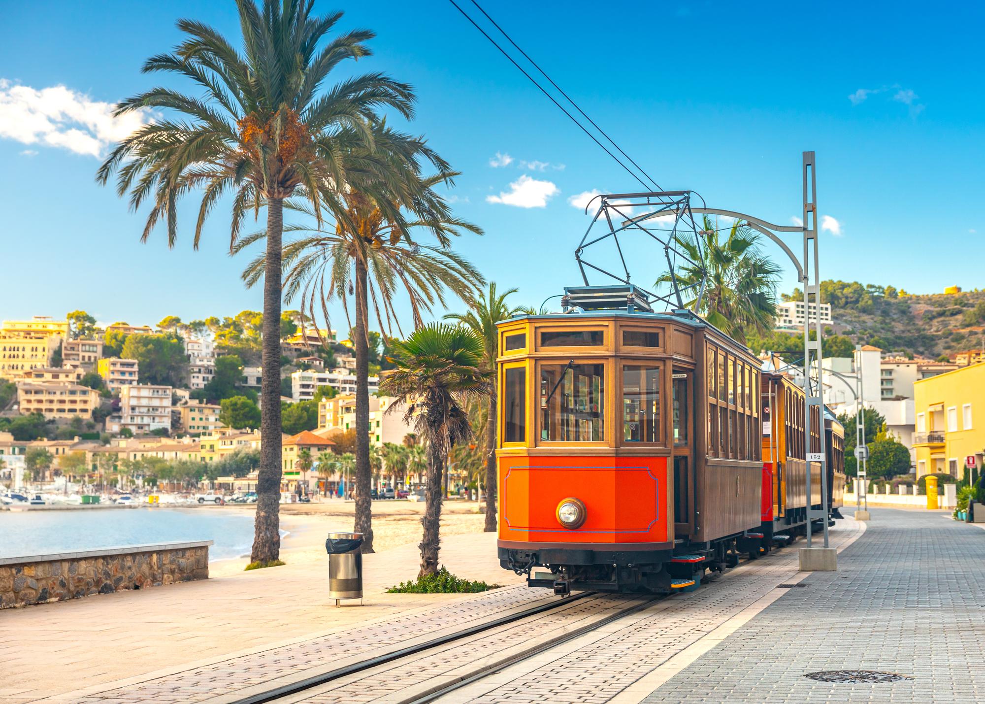 Experience the scenic beauty of Puerto Soller in Spain's Balearic Islands, with its picturesque beach, tranquil sea, and iconic old orange tram transport | Beach, scenic, sea, transport, old, Puerto Soller, Balearic Islands, Spain | Fraser