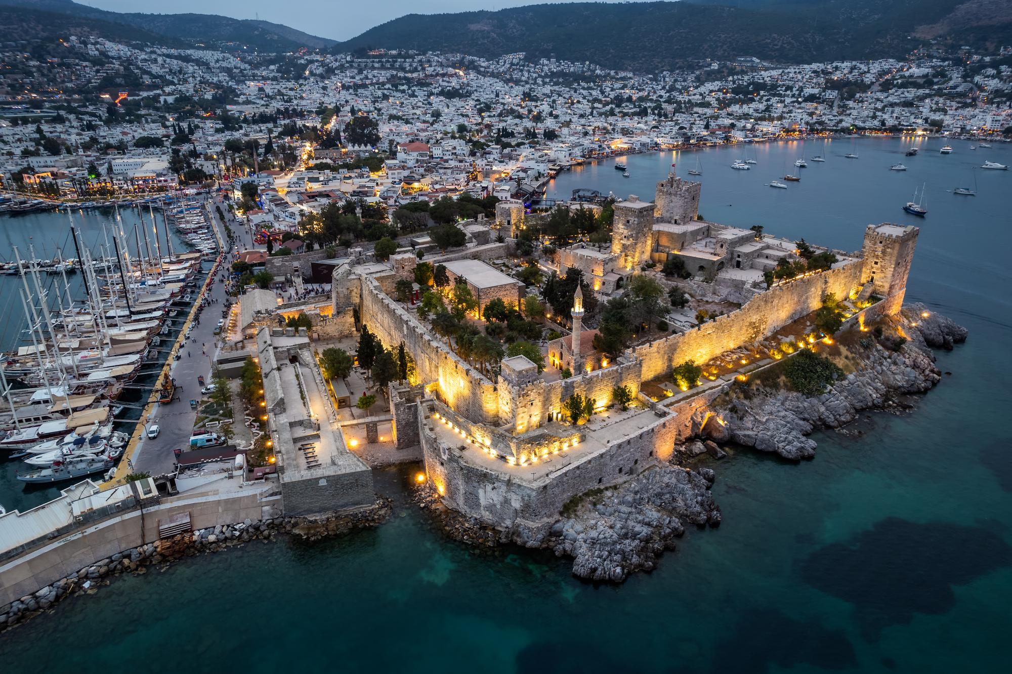 Take in the aerial view of Bodrum Castle overlooking the harbor and marina, with the picturesque coastline illuminated in the evening light | Aerial view, harbor, marina, coastline, evening, Bodrum, Turkey | Fraser